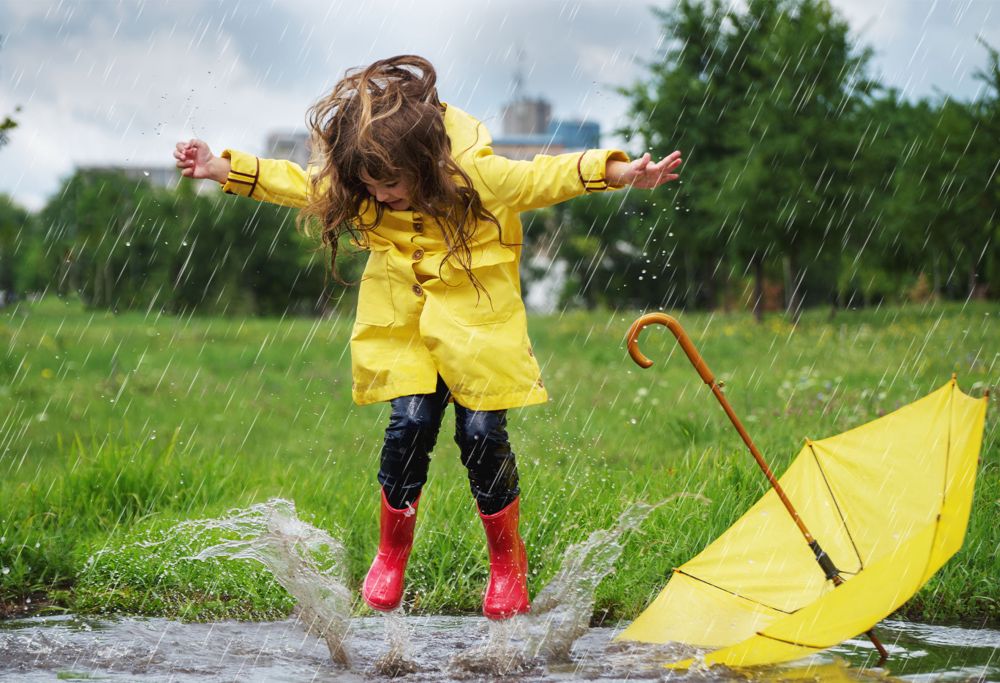 A child in a raincoat and boots splashes in a puddle of water. 