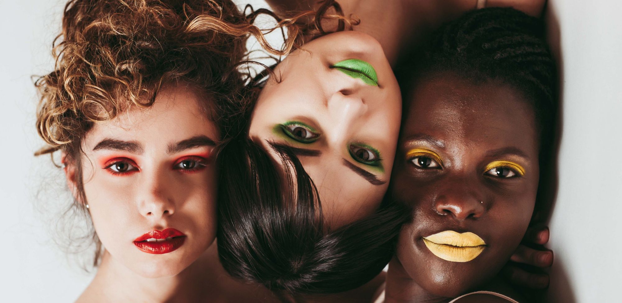 Three people wearing vibrant colored lipstick and eyeshadow pose together. One wears red, one green and one yellow.  