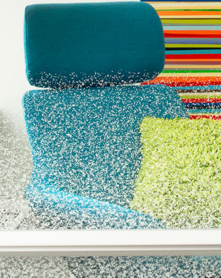 Blue lounge chair with a bright green pillow, partially obscured by textured frosted glass, with a colorful striped artwork on the wall behind it. 