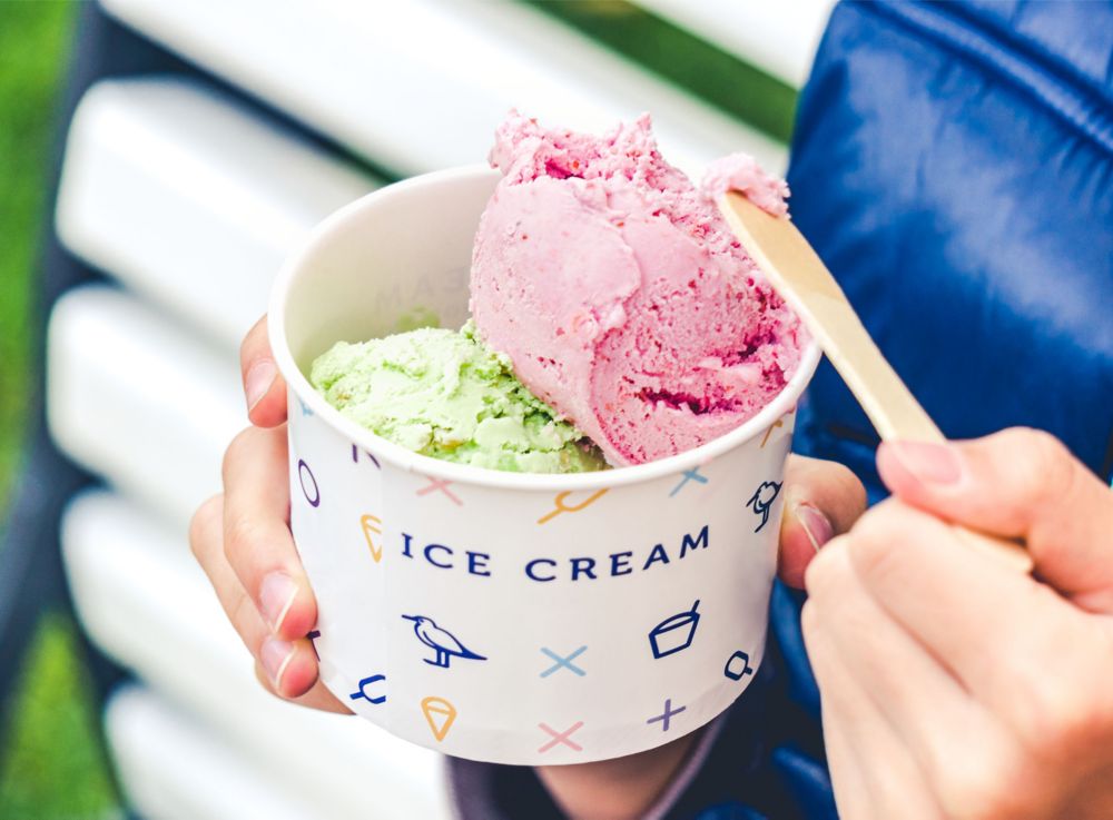 A person eats ice cream out of a paper cup with a wooden spoon. 