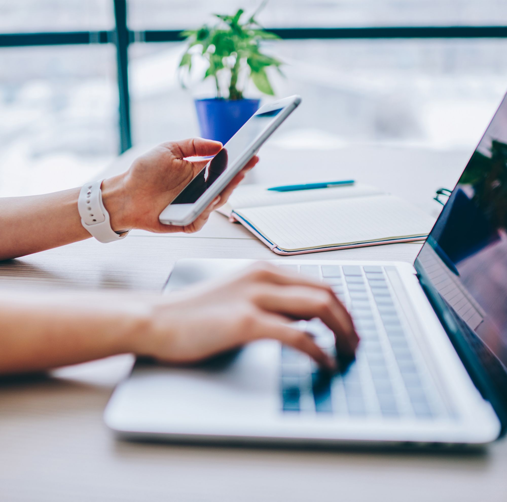 A person holds a cell phone in their left hand and types on a laptop with their right. 