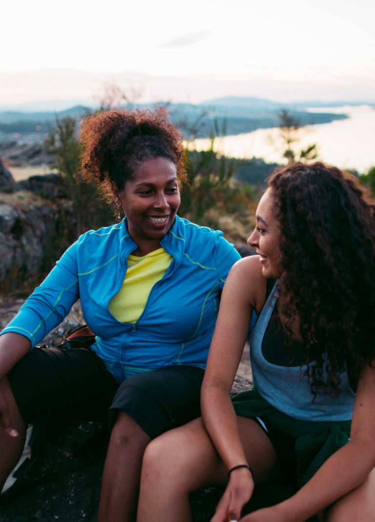 Two women sit and talk at the top of a hill overlooking a lake. 