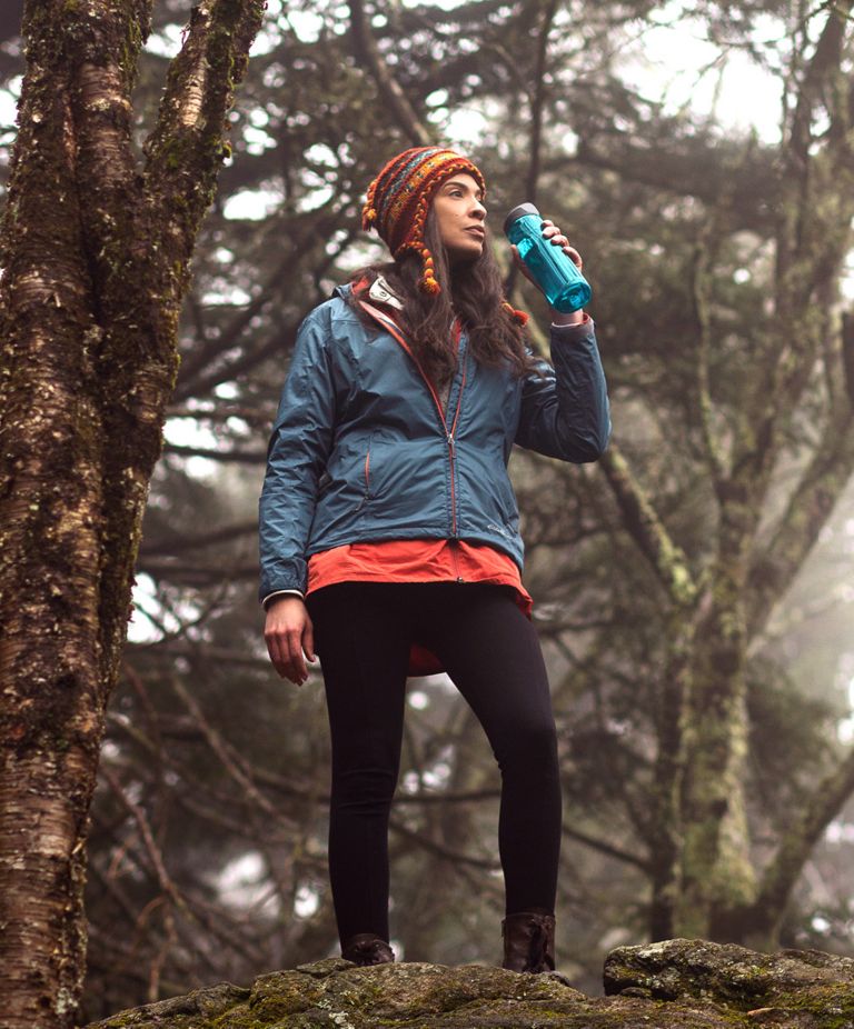 A hiker in the forest ready to drink water from a dark blue, reusable bottle made of Tritan. 