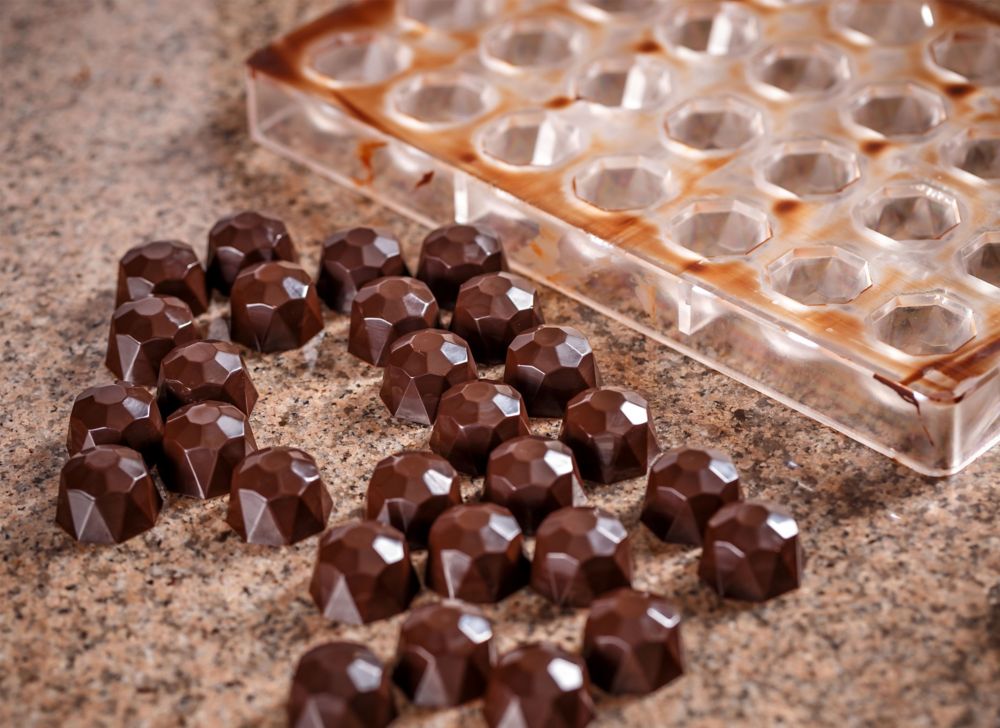 Numerous small chocolate candies sit on a counter beside a used mold. 