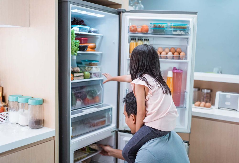 A father grabs food from the freezer as his child sits on his shoulders. 