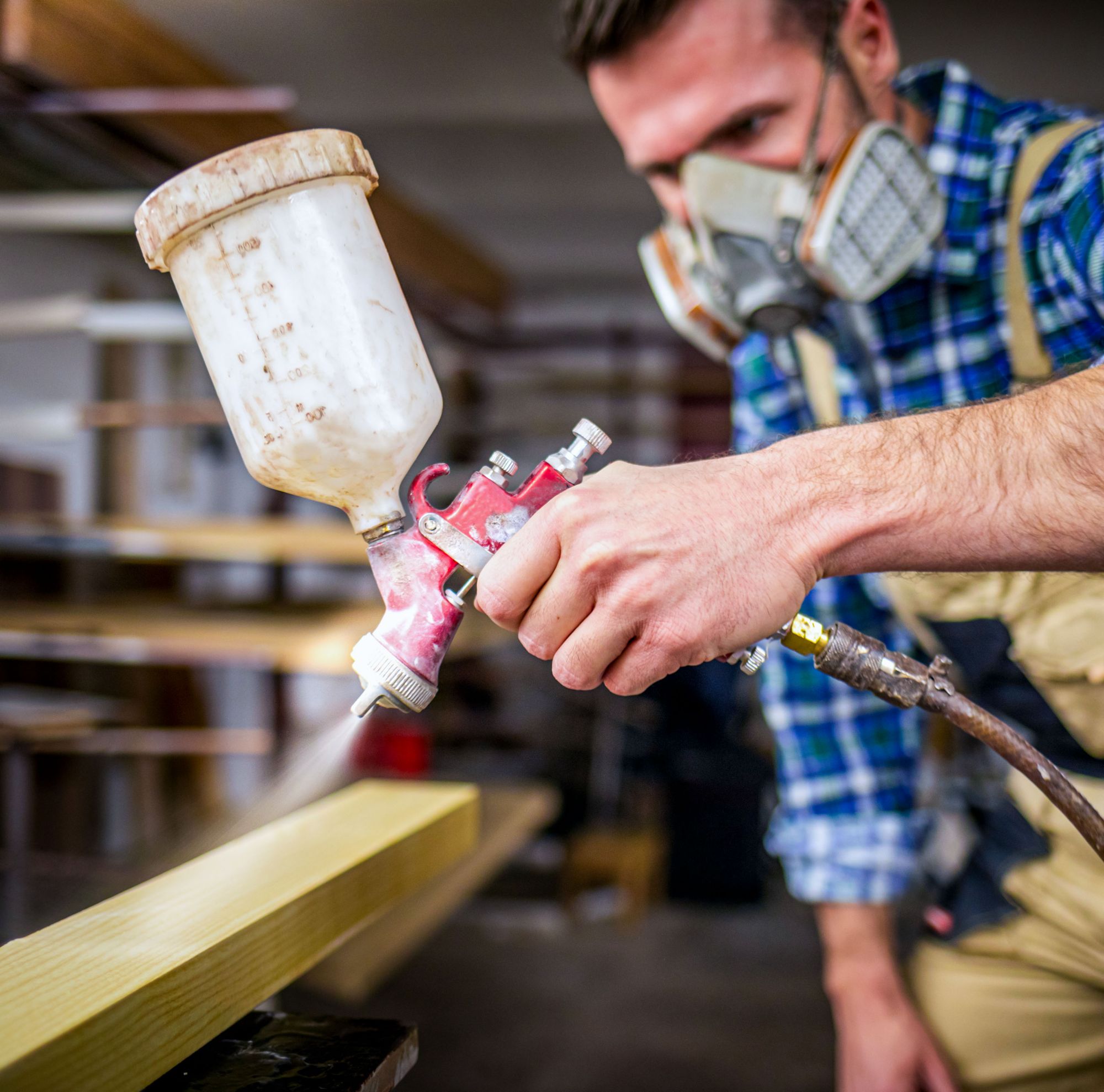 A woodworker in PPE sprays a coating onto a wood board. 