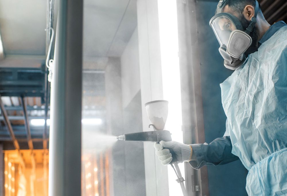A man in full PPE, including a hazmat suit and face shield, sprays coatings onto metal. 