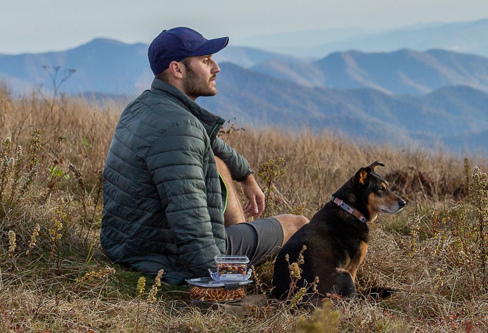 A man and dog sitting on the ground in the mountains with two plastic food containers. 