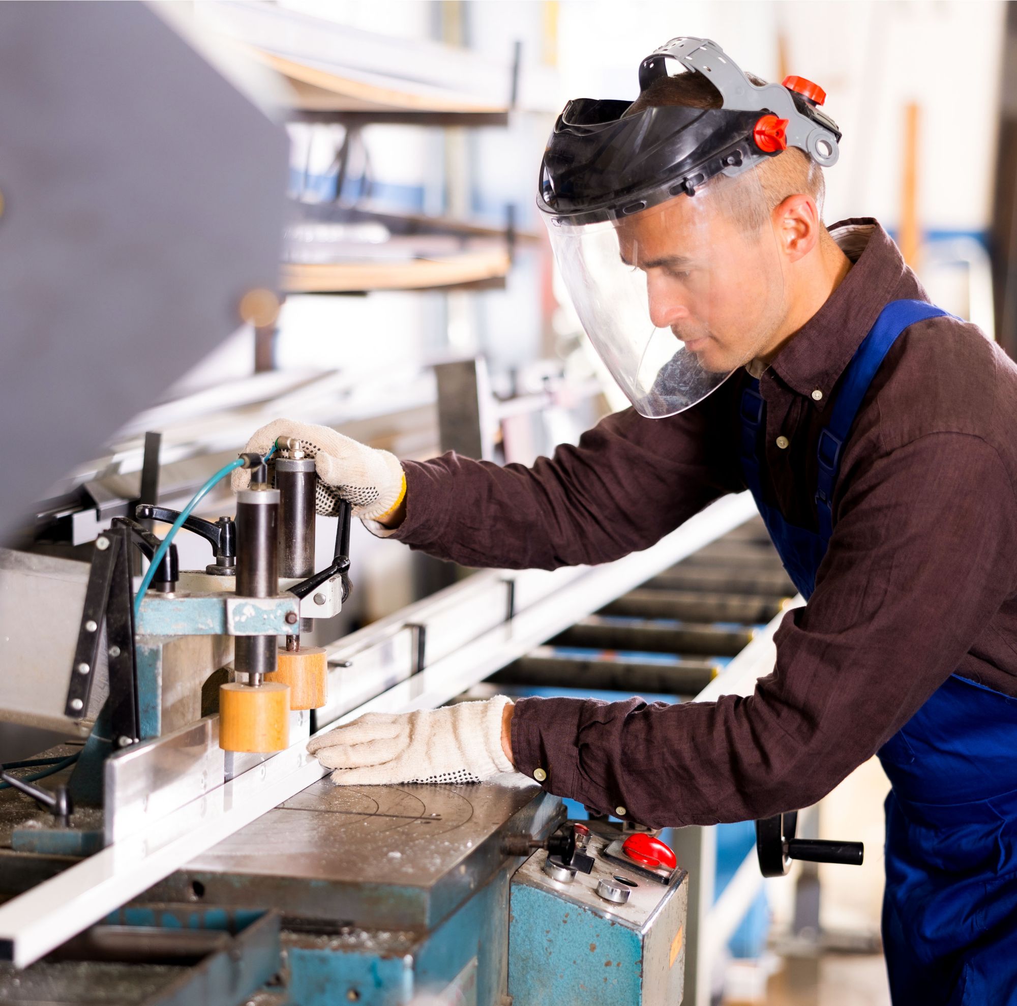 A man in a large, clear, full-face shield operates a cutting machine. 