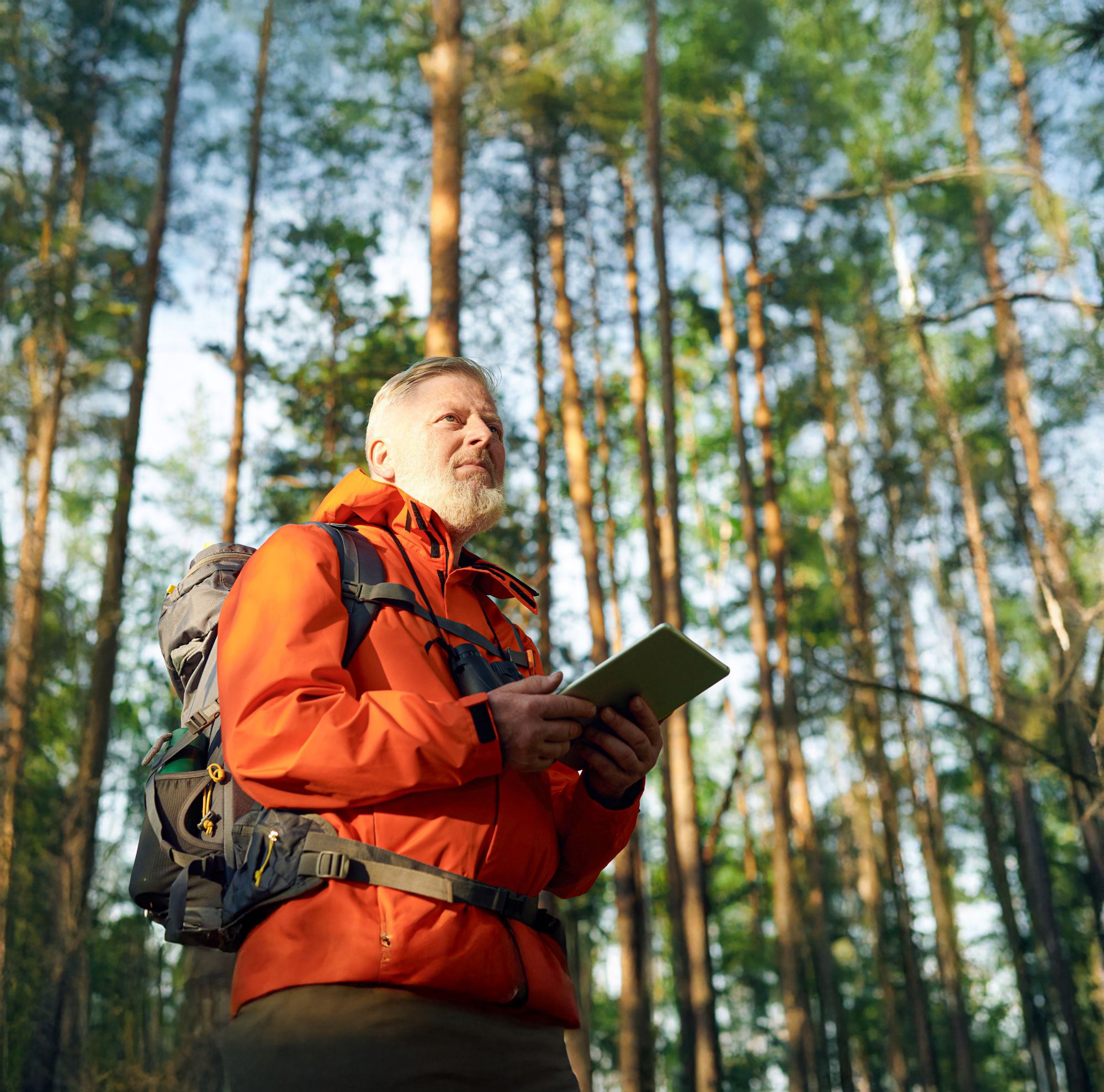 A man in an orange rain jacket looks out at a forest while holding a tablet. 
