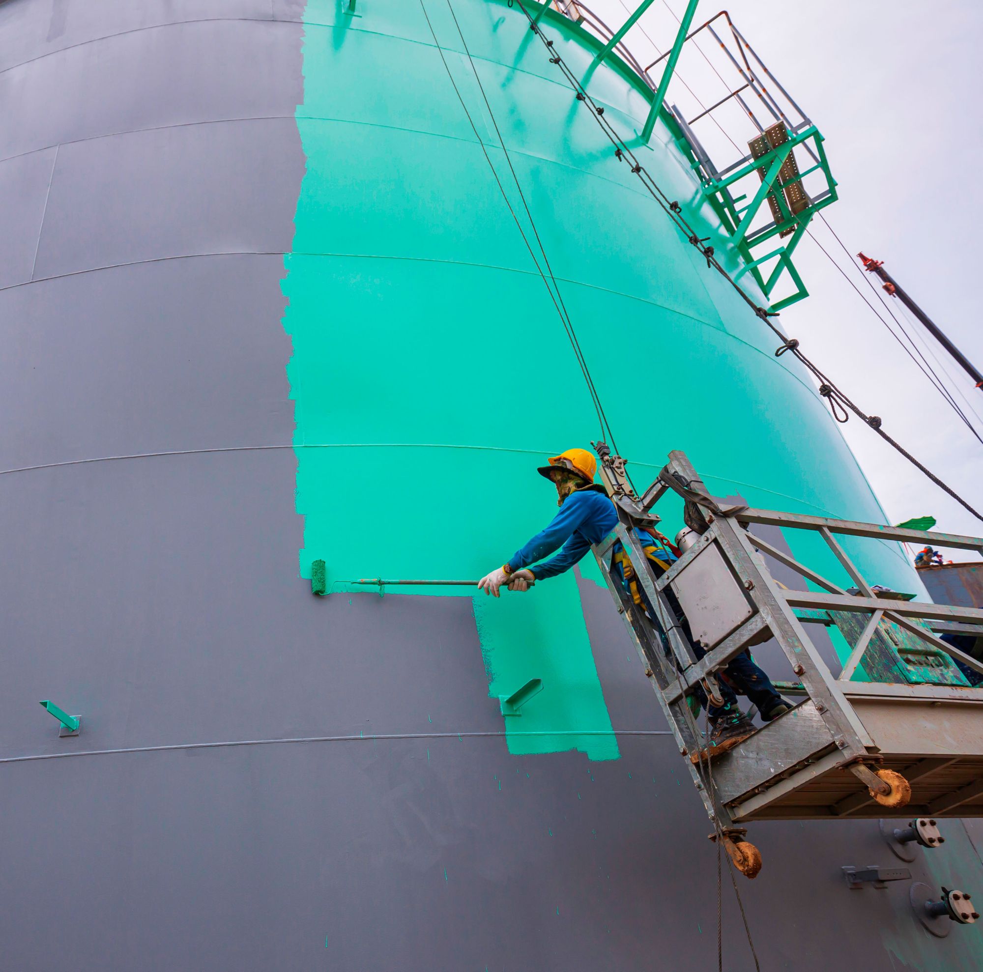 Painter applying green paint to the side of a large metal container. 