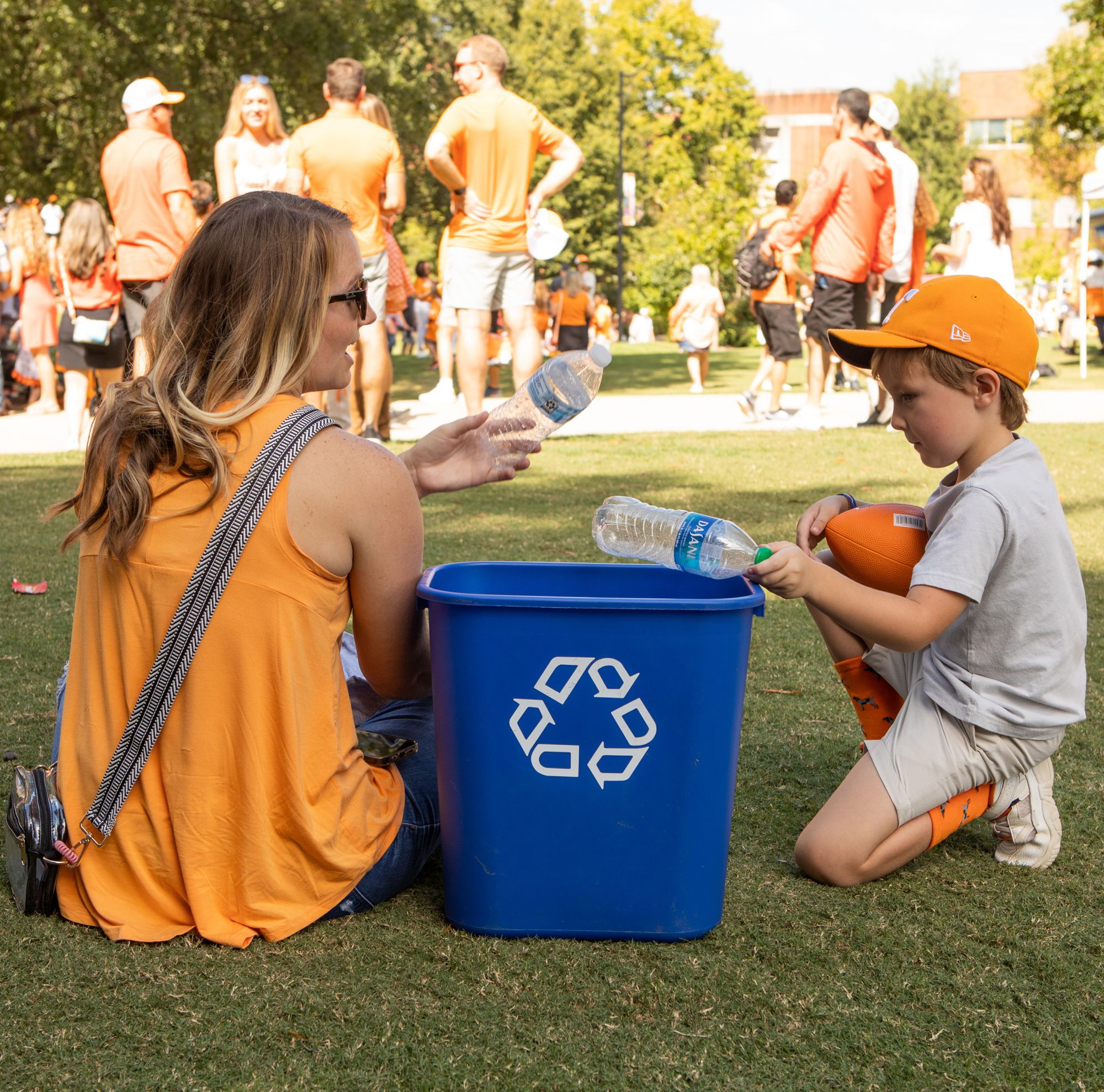 A woman and a child in a ballcap toss plastic bottles into a recycling bin. 