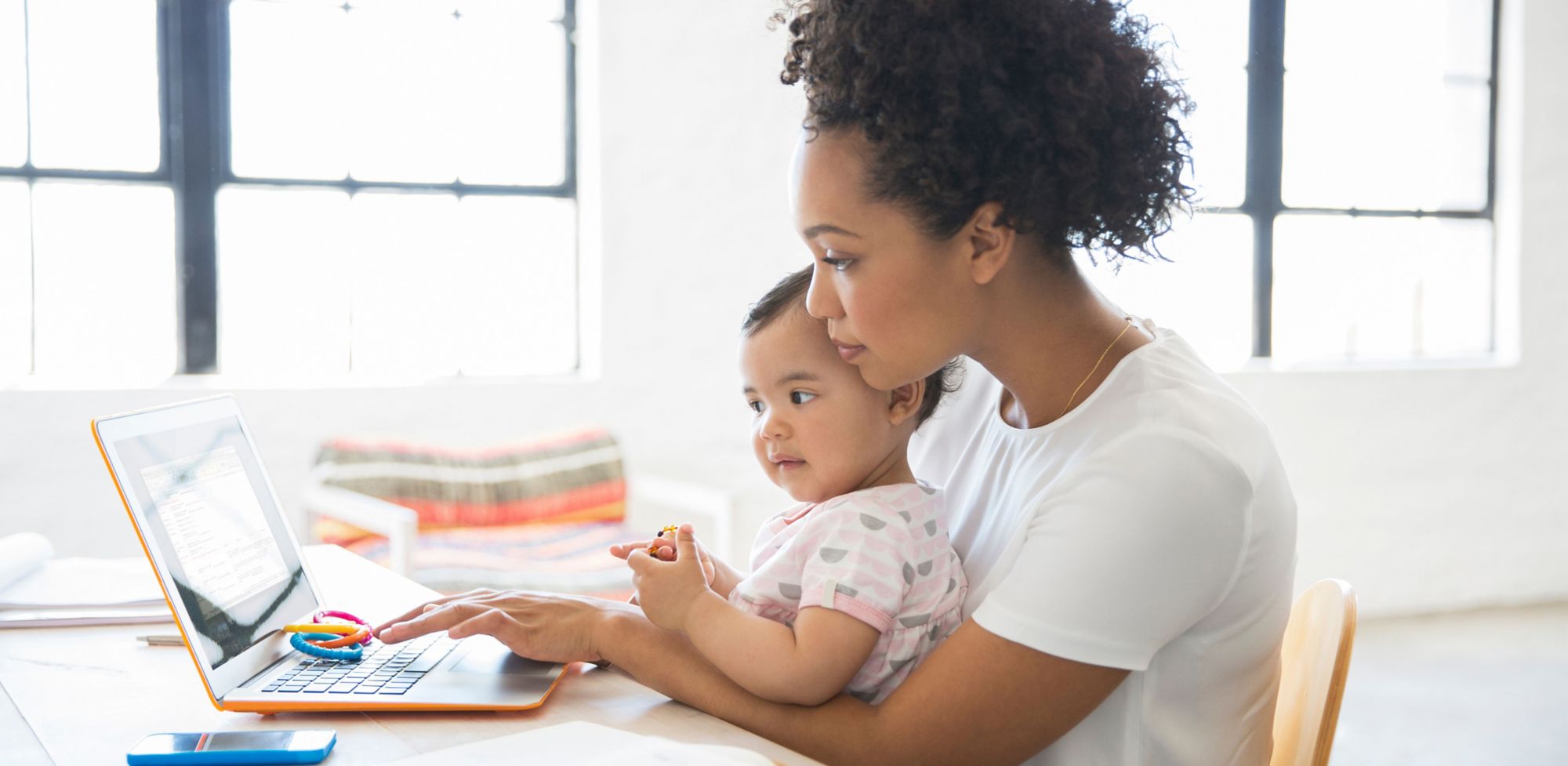 Una madre se sienta con un bebé en su regazo mientras usa su computadora portátil. 