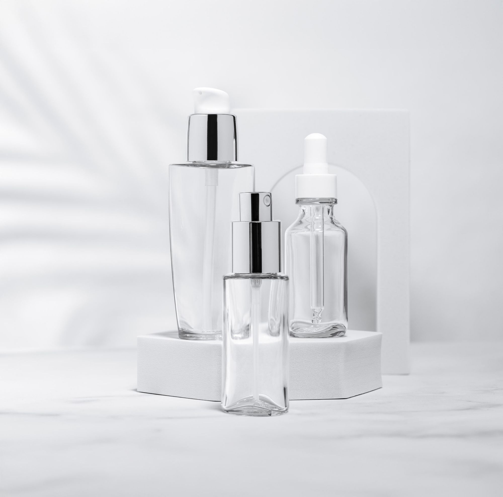 Clear cosmetic bottles with pumps and droppers displayed on white stands against a soft, neutral background. 
