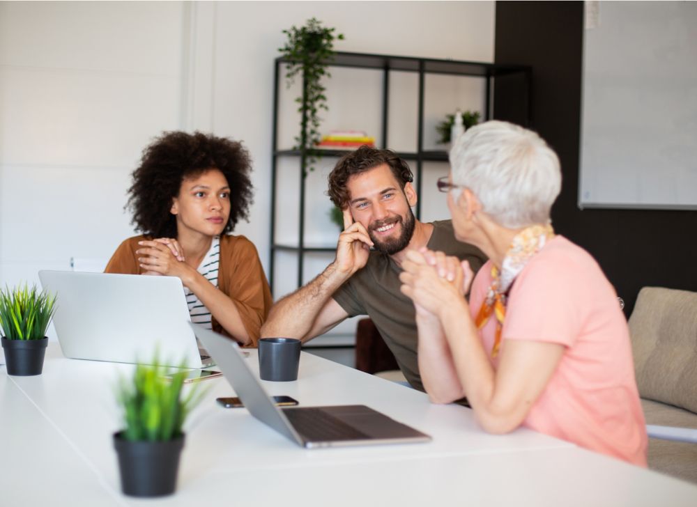 Three people working on their laptops talk in a conference room. 