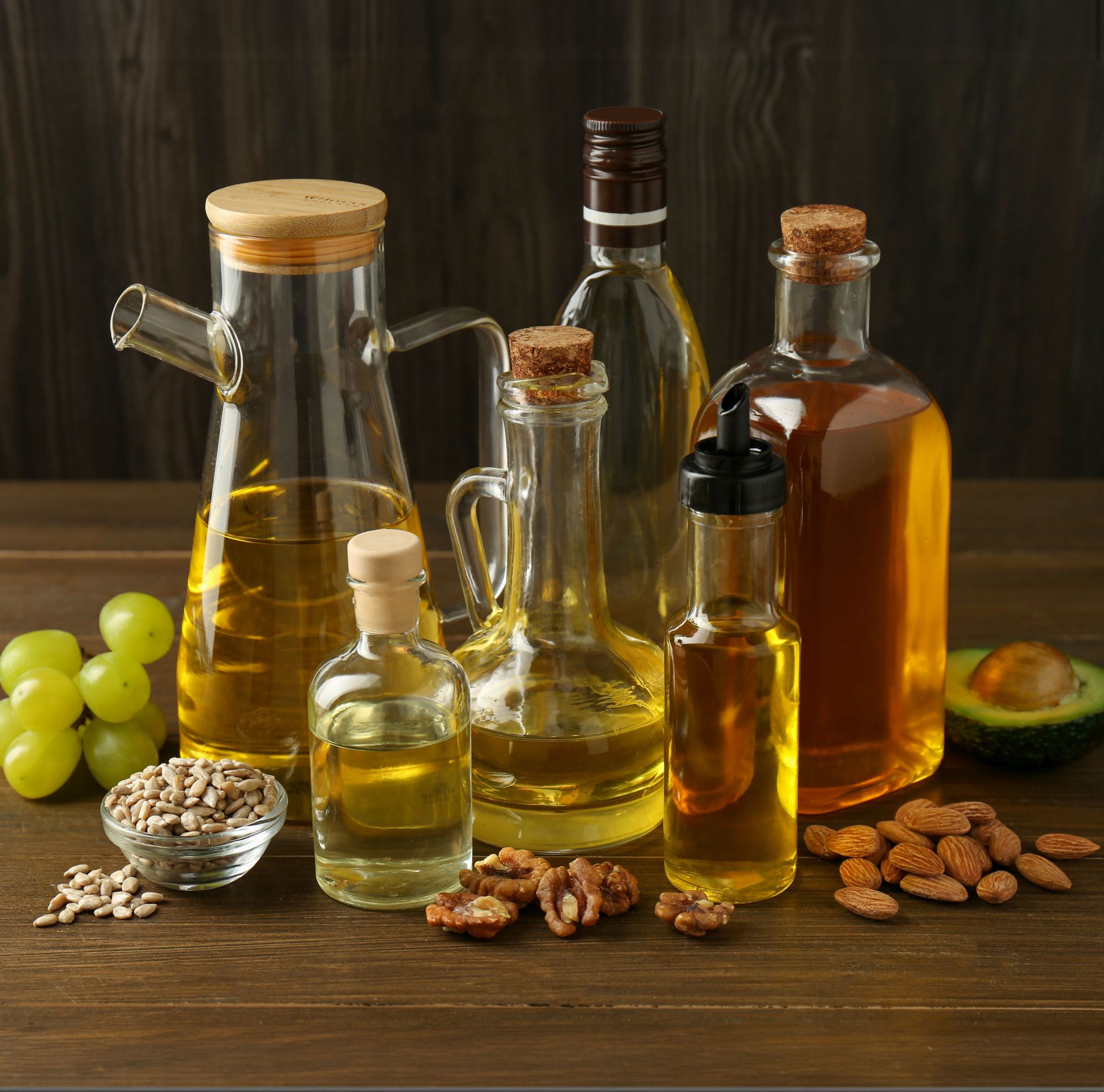 Six bottles of cooking oils surrounded by various nuts and grapes. 