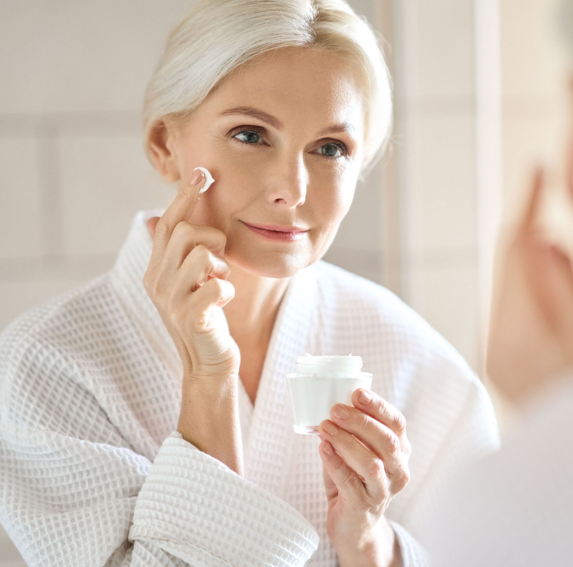 Woman in a white robe applies face cream. 