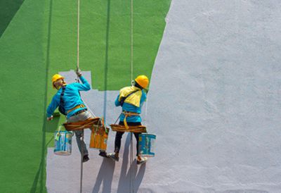 Two people are suspended above the ground, sitting on small surfaces as they paint an exterior wall green. 