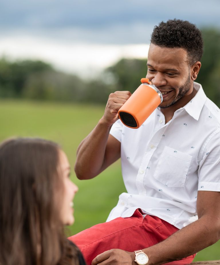 A person smiling as he drinks from an orange tumbler cup with a handle. 