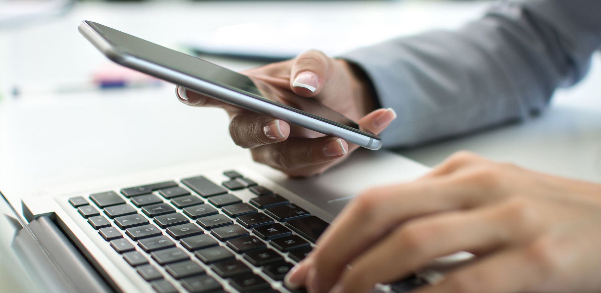 A person is holding a cell phone while typing on a laptop. 