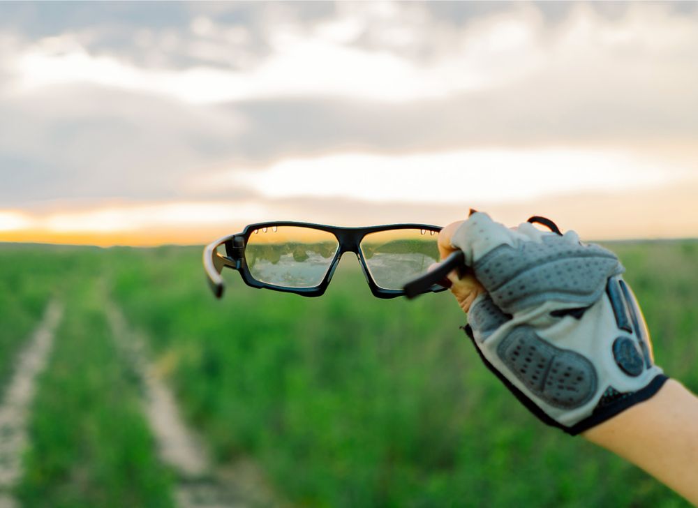 A person wearing cycling gloves holds up a pair of sports glasses. 