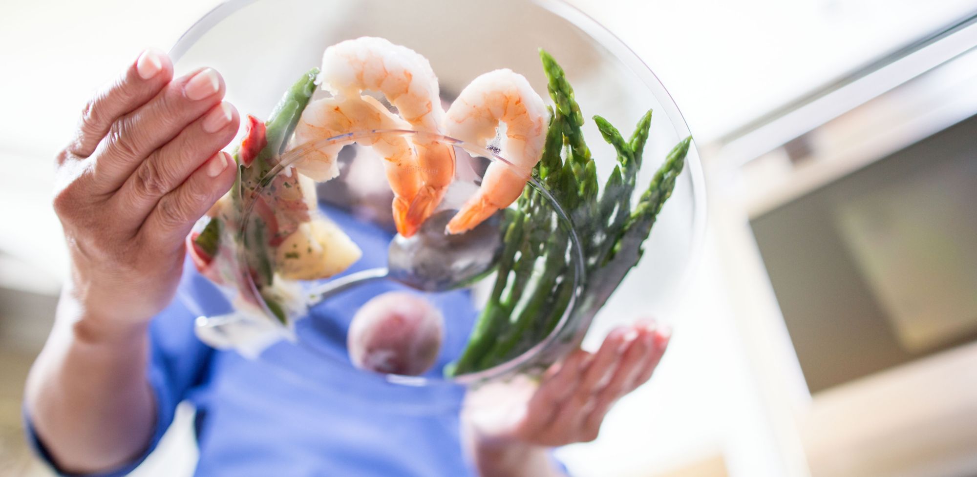 A person holds a clear bowl with shrimp, asparagus and potato salad. 