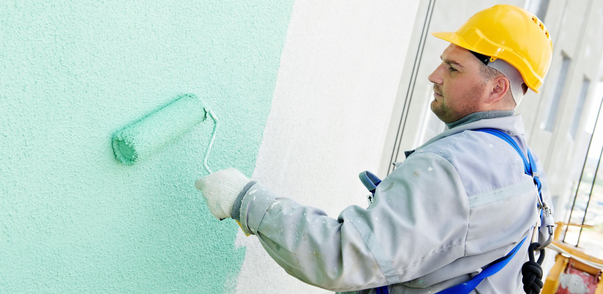 A man using a roller applies bluish-green paint to a wall. 