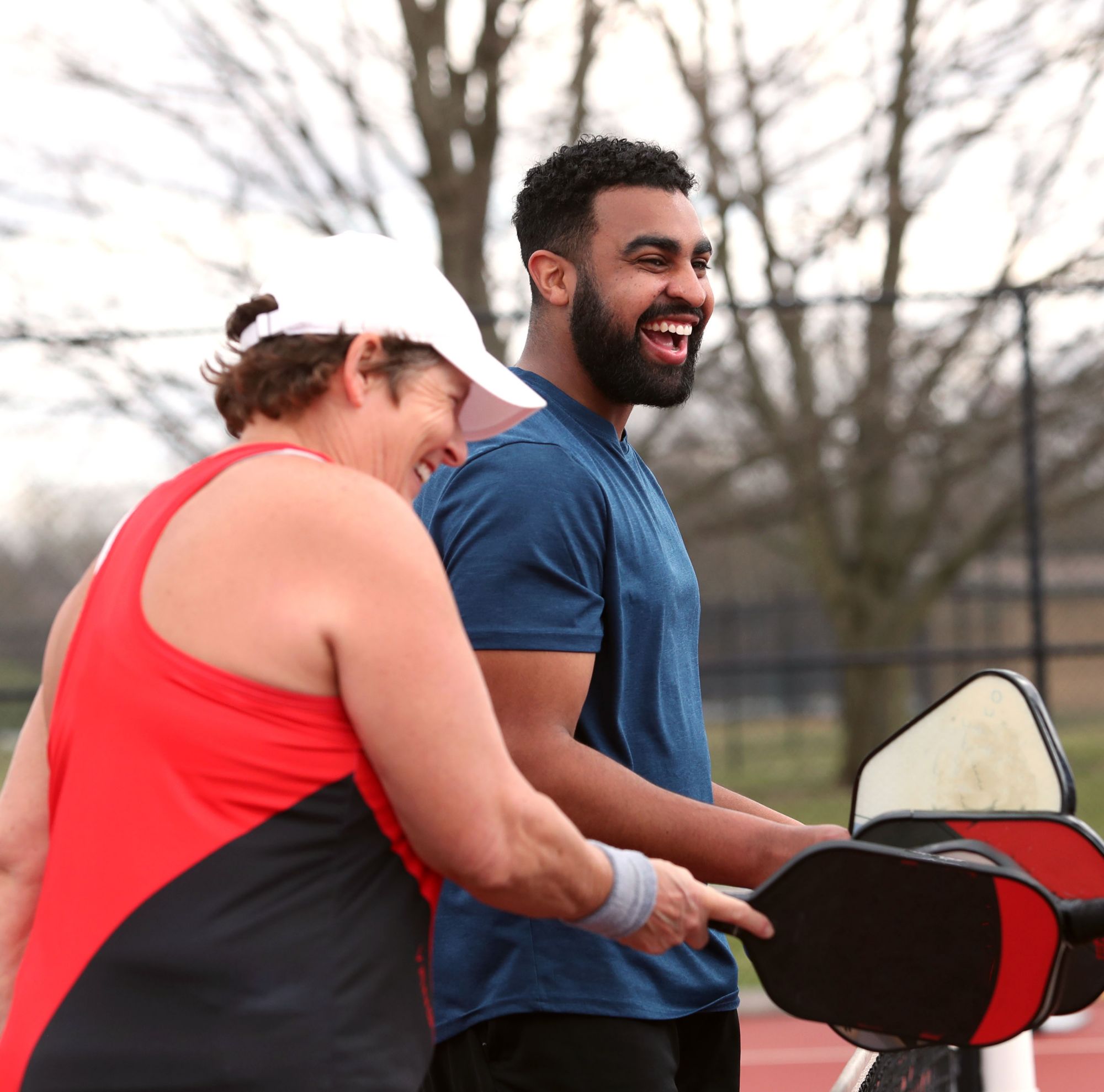 Two people laugh with pickleball paddles outside. 