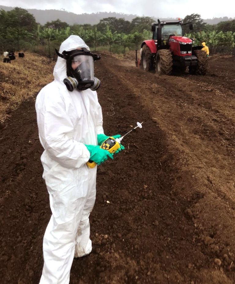 Person in white hazmat suit, full-face respirator mask, and green gloves holding a soil testing instrument in a field 