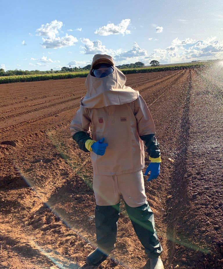 A farmer dressed in full PPE, including eye protection, gloves and boots, standing in a plowed field 