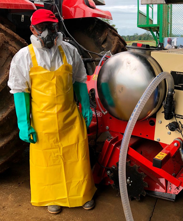 Person in white suit, yellow apron, half-face respirator mask, protective glasses and green gloves next to a soil injector machine. 