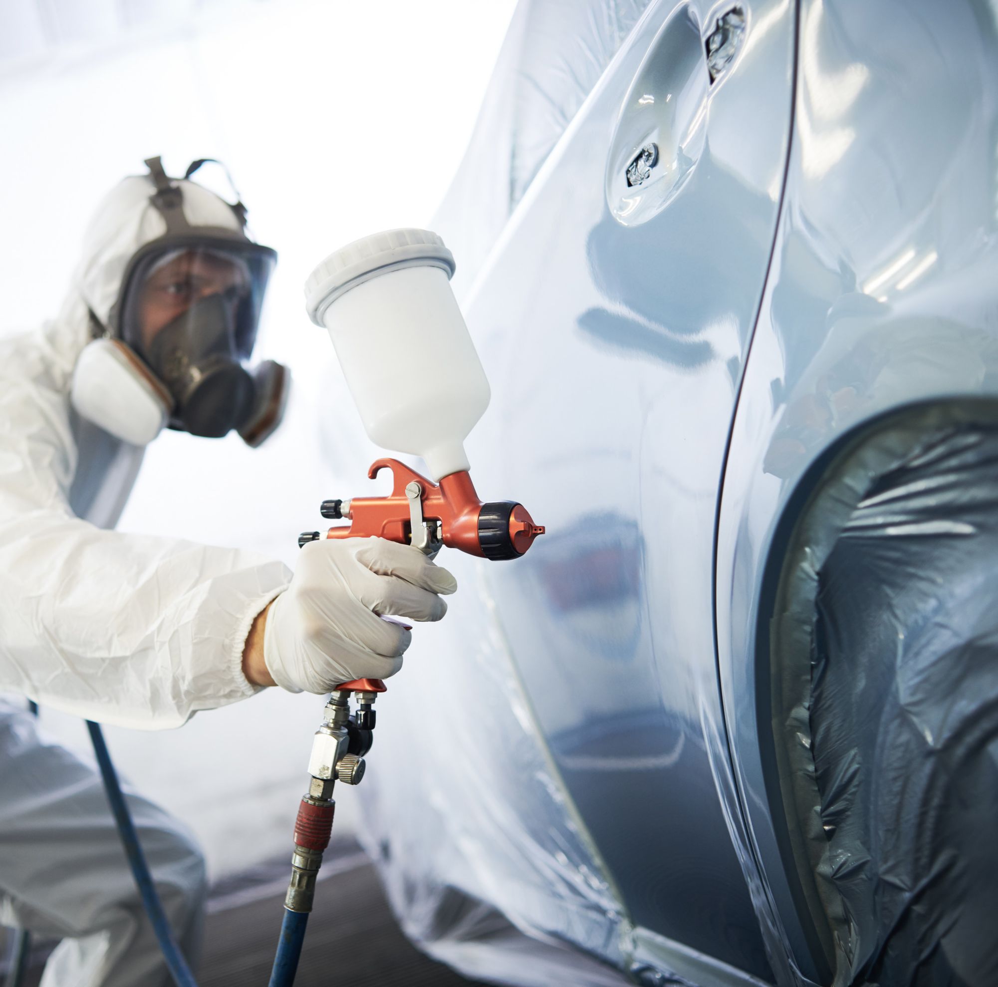 A man in white protective gear sprays gray paint onto the rear driver-side door of a car. 
