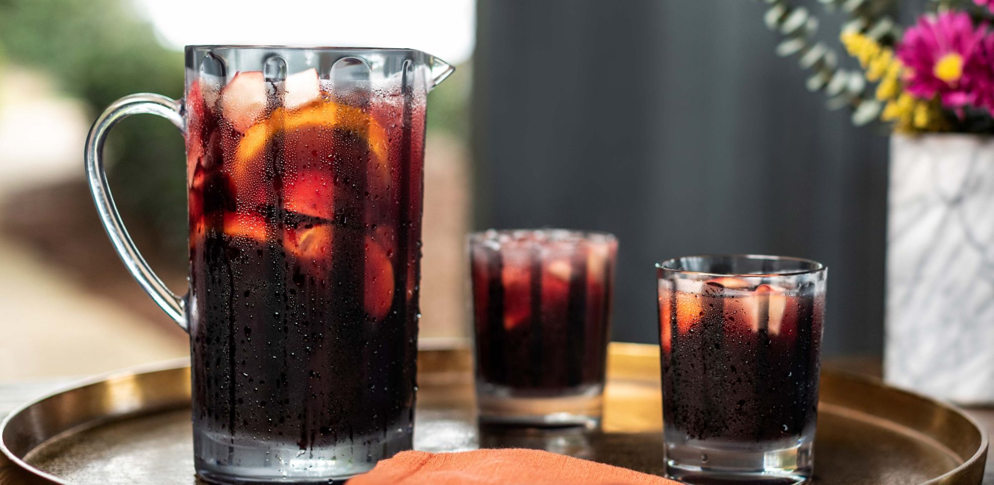 A clear pitcher and two clear cups filled with ice and red sangria. 