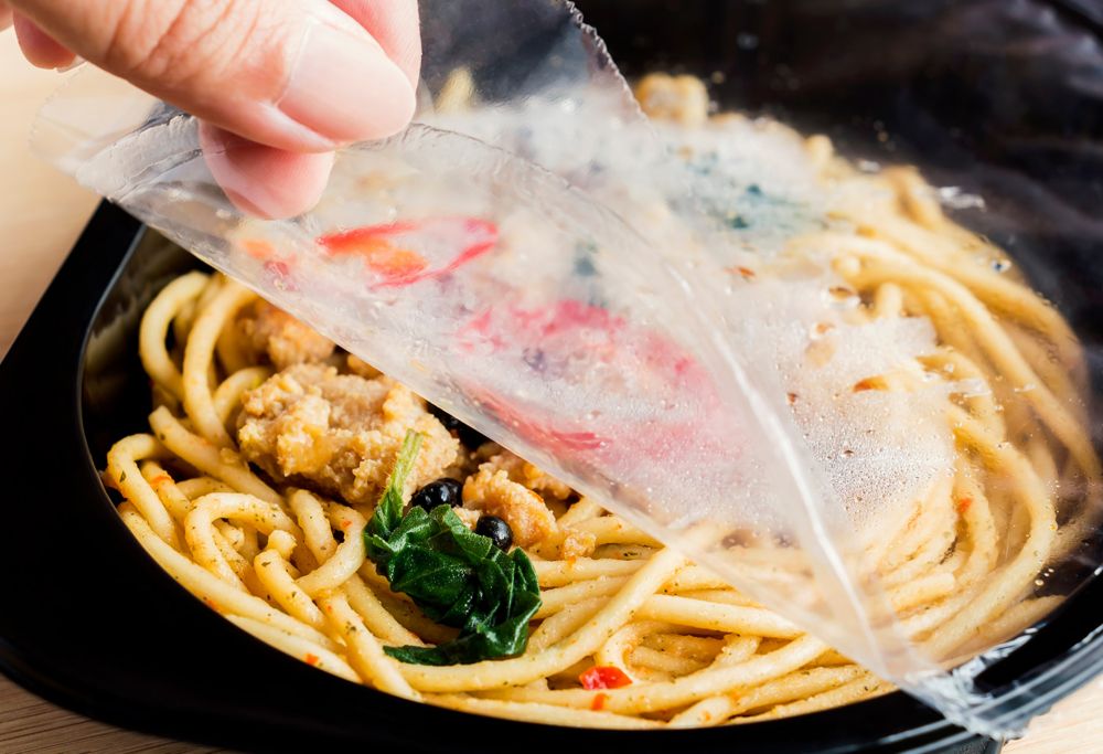 A person lifts plastic film off a frozen noodle meal. 
