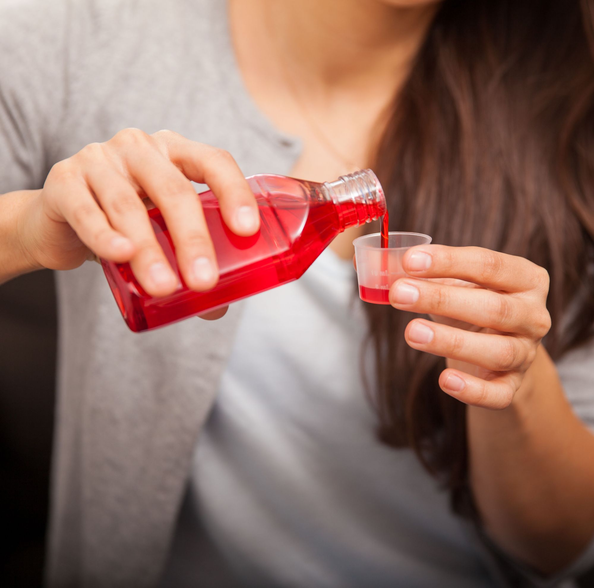 A person pouring red cough syrup into a medical measuring cup. 