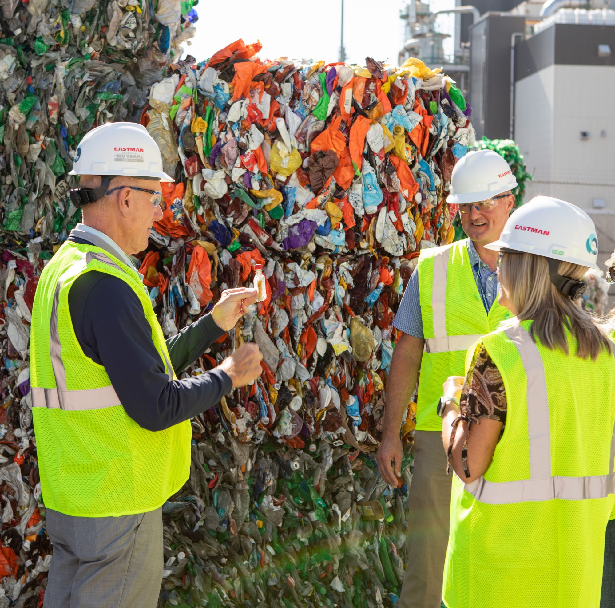 Three people in safety vests and Eastman hard hats stand near bales of plastic. 