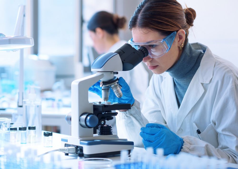 A person in gloves and goggles looks into a microscope.  