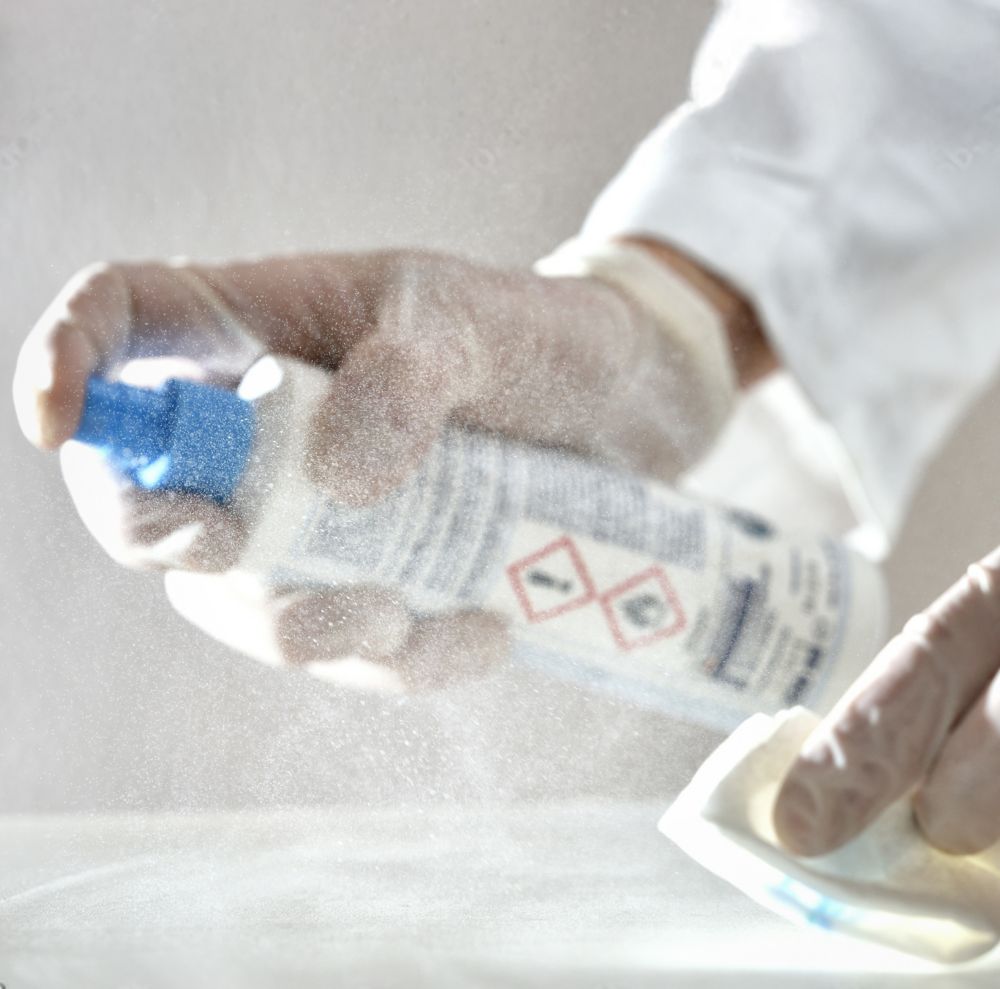 A person wearing gloves and a lab coat sprays and wipes a surface clean.  