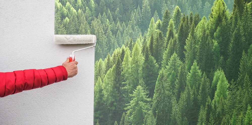 A person rolls paint onto a wall. On the wall is a partially done landscape full of evergreen trees. 