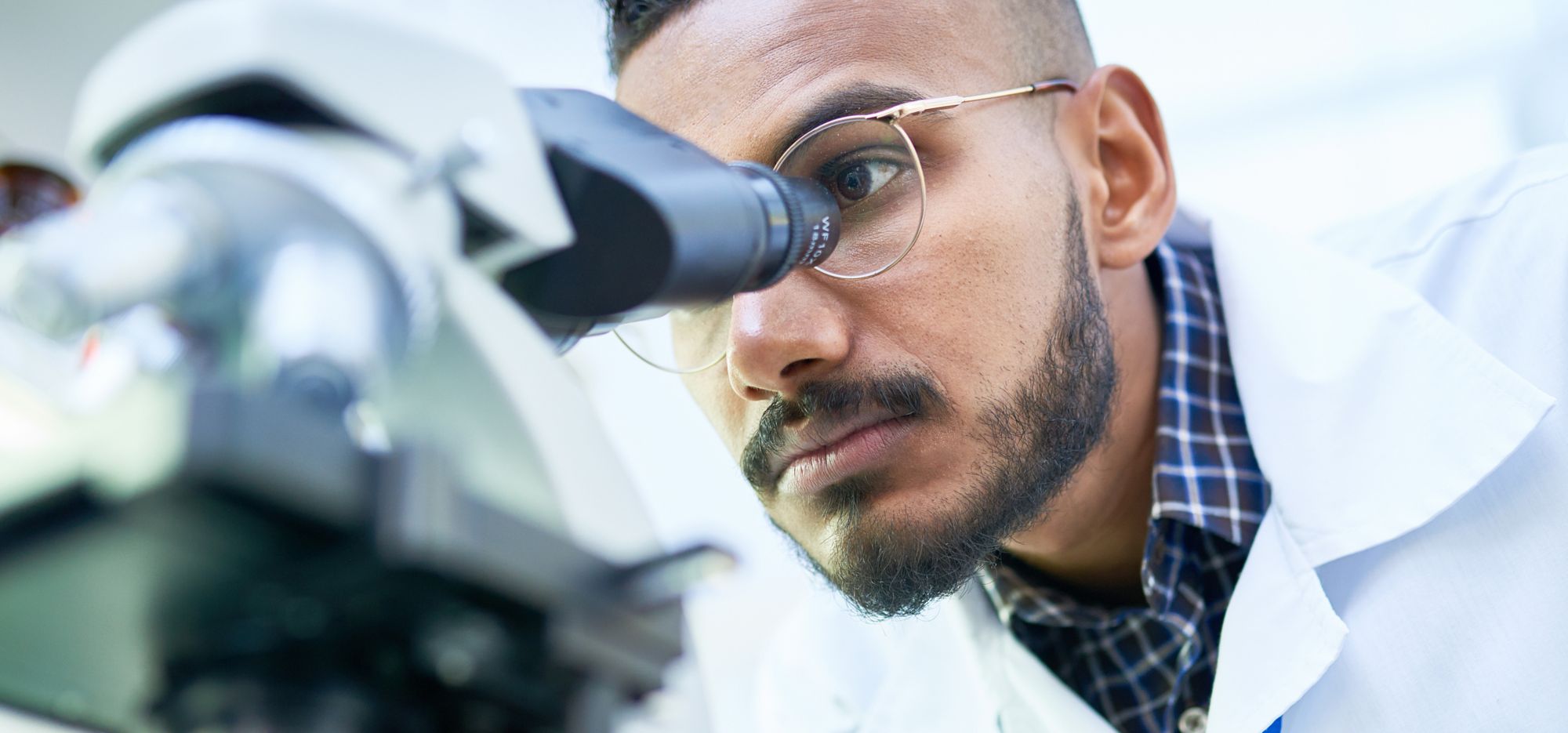 A person in a lab coat looks into a microscope. 