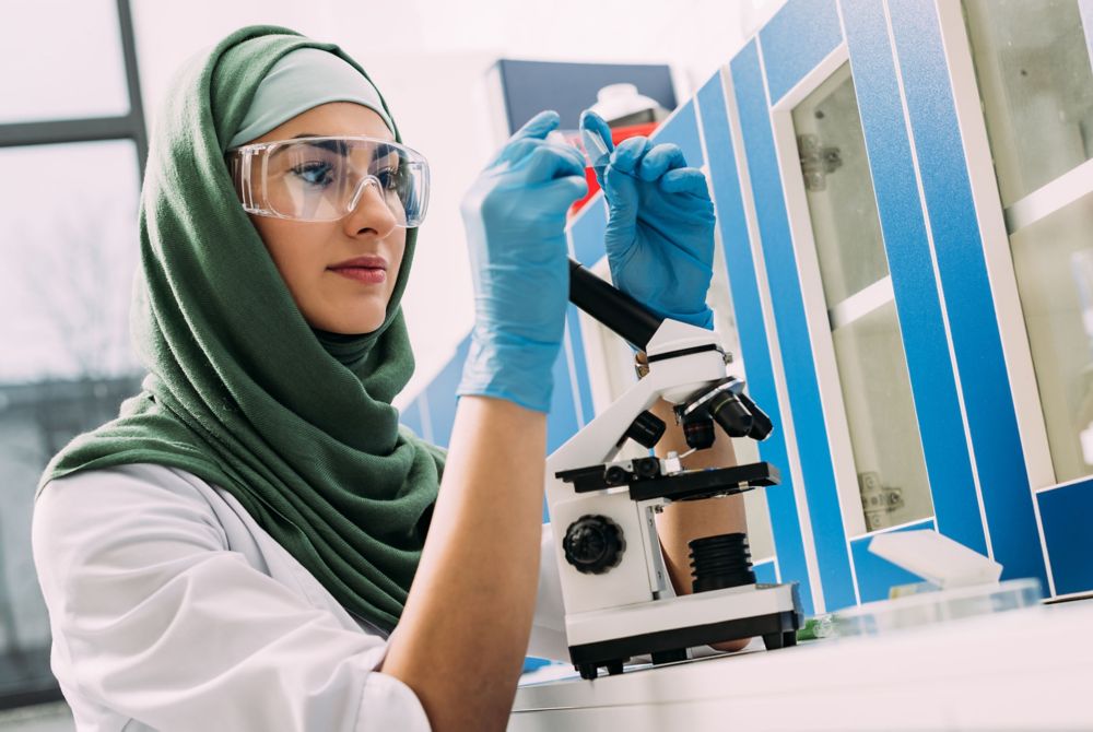 Female scientist wearing hijab, goggles and gloves observing lab sample. 