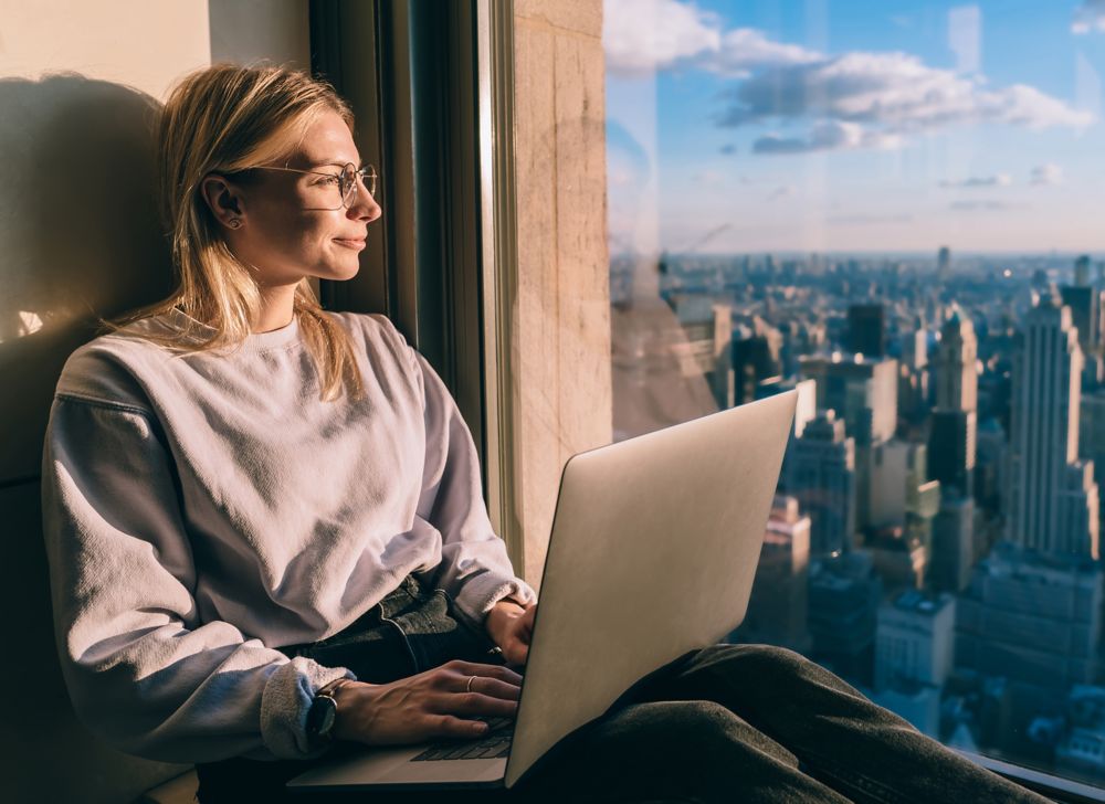 A person sitting with laptop in hand and looking out the window of a skyscraper. 