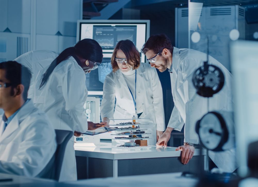 Three technicians in white lab coats looking at data on a tablet. 