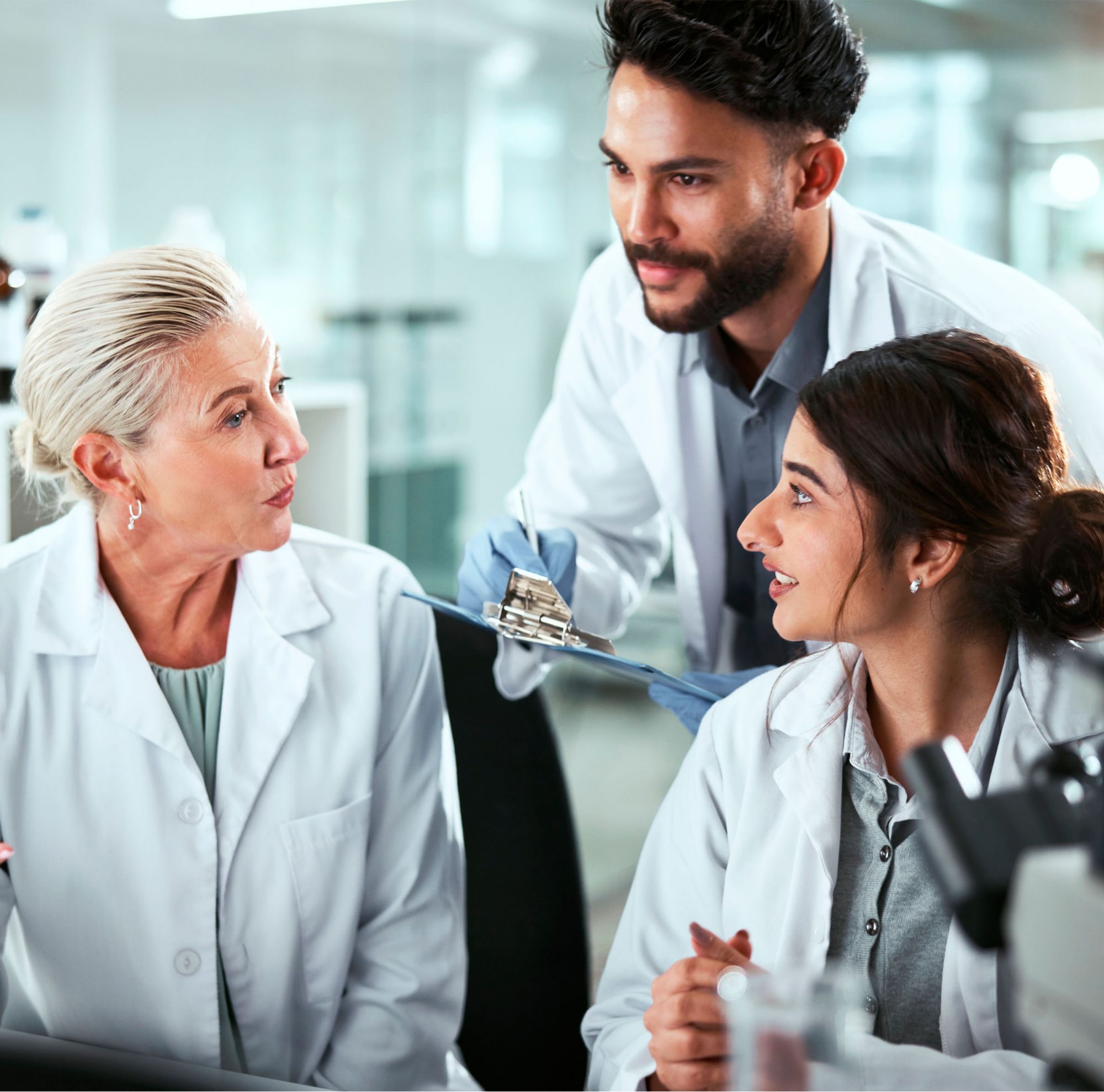 Three scientists talk to each other, surrounded by microscopes and computers. 