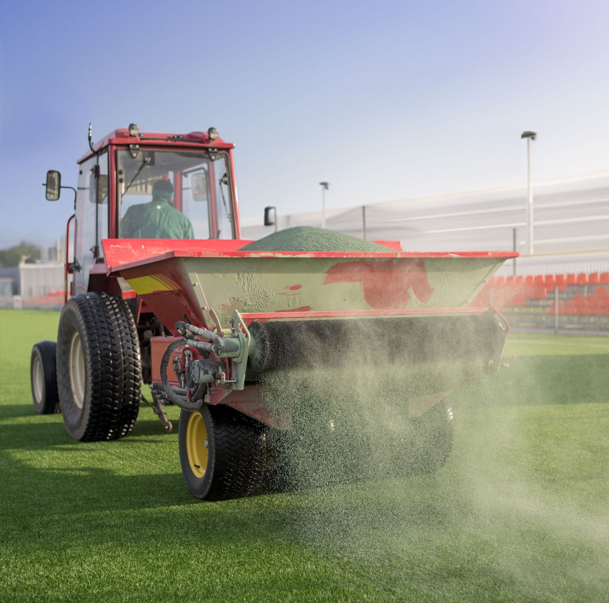 A tractor driver is spreading sand on an artificial grass footfield. 