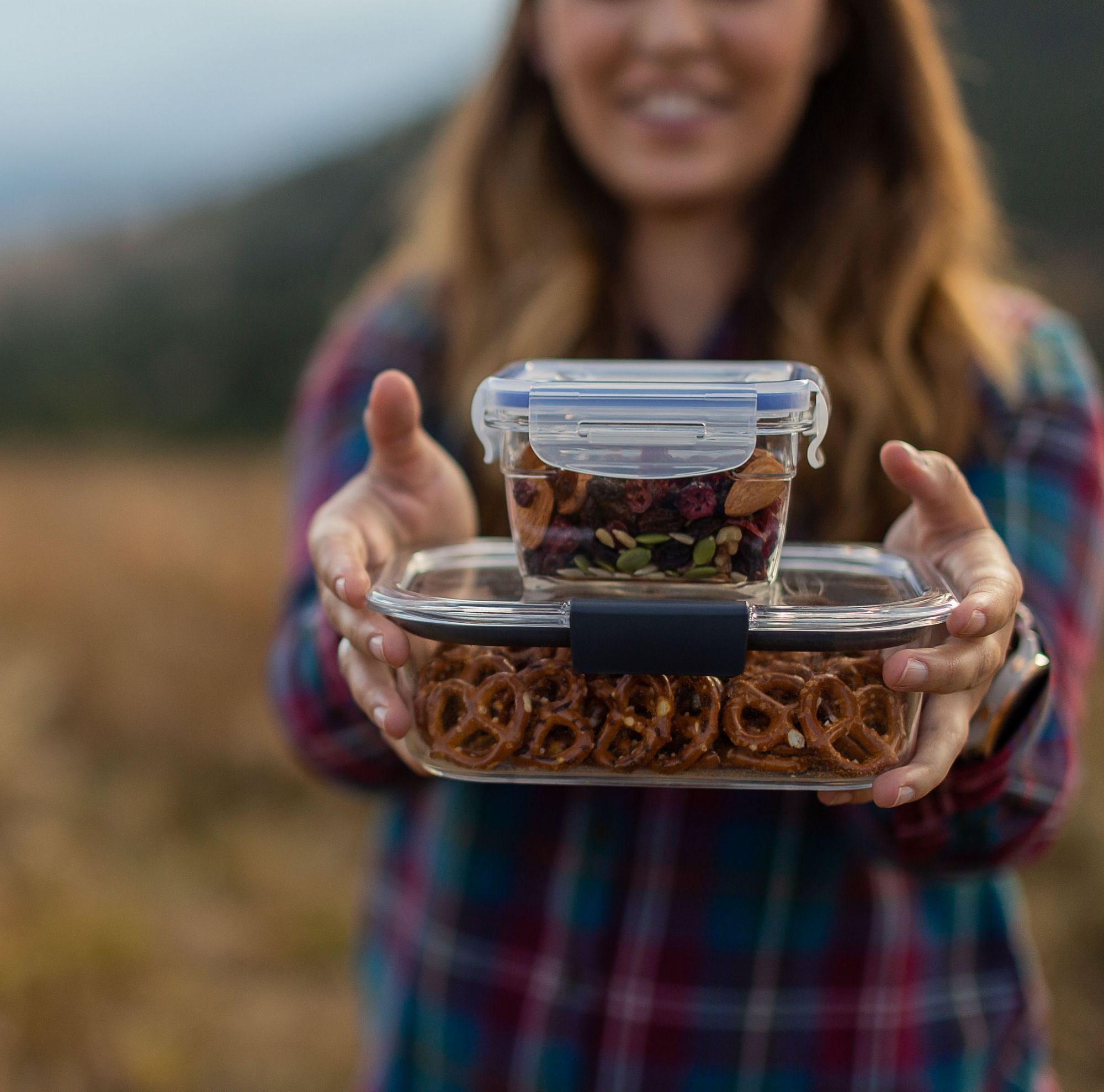A person holding two food containers, one filled with pretzels and the other with trail mix.  