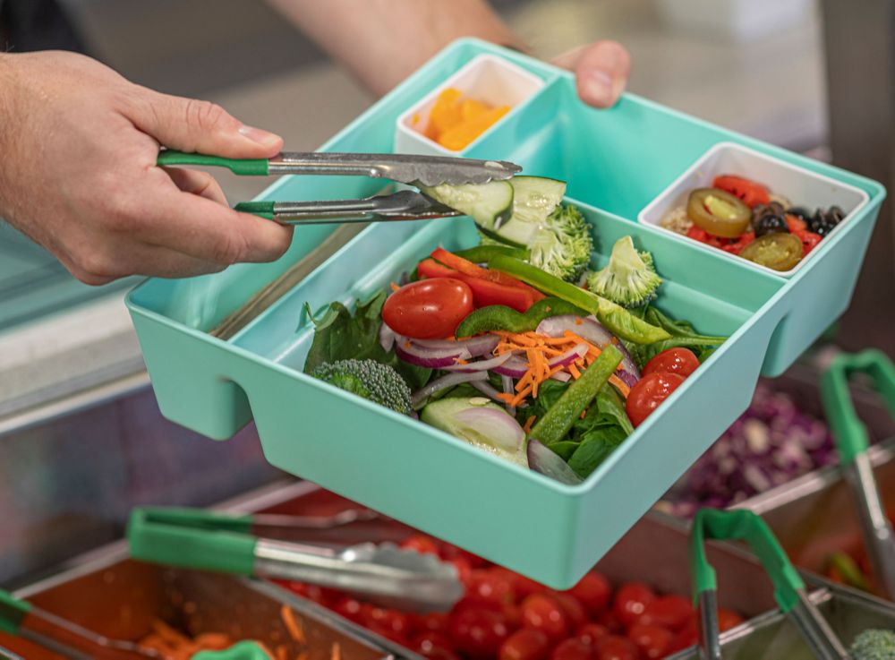 A person adds cucumbers to a salad in blue bento box made from Tritan Renew. 