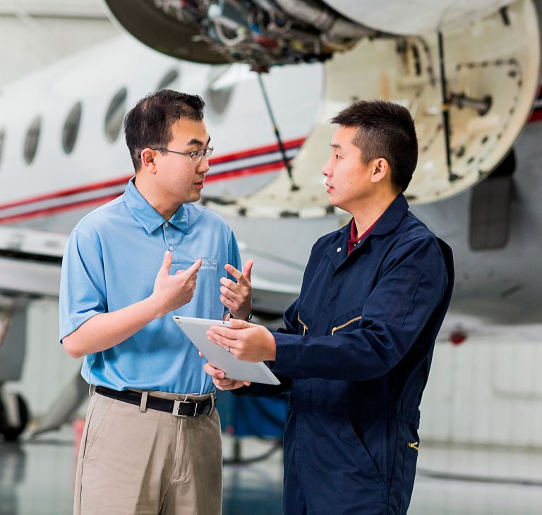 Two men talk while looking at a paper and standing in front of a plane in a hangar. 