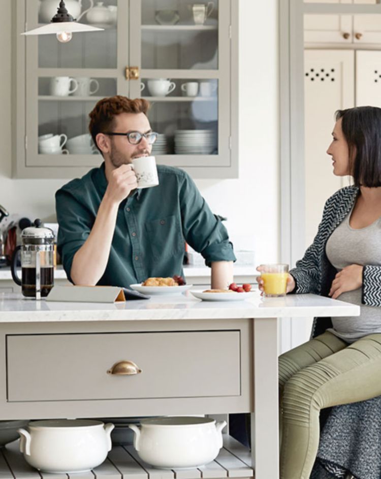 A modern kitchen with a gray island, glass‑front cabinets, and large windows, with two people seated at the island having breakfast. 