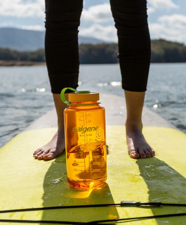 A person stands on a surfboard that holds a Tritan Nalgene bottle. 